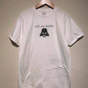 Op de afbeelding: Wit T-shirt met een zwarte Darth Vader-afbeelding en de tekst "call me daddy" gedrukt op de voorkant.
