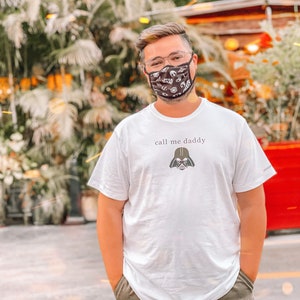 Op de afbeelding: Een wit T-shirt met de tekst "call me daddy" en een zwarte Darth Vader afbeelding. De persoon die het shirt draagt, draagt ook een zwart mondkapje met een wit patroon.
