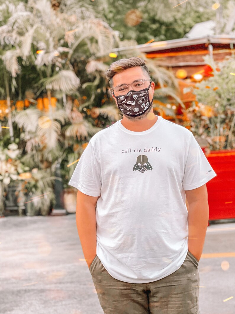 Op de afbeelding: Een wit T-shirt met de tekst "call me daddy" en een zwarte Darth Vader afbeelding. De persoon die het shirt draagt, draagt ook een zwart mondkapje met een wit patroon.