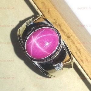 Peut inclure: Une bague en argent avec une grande pierre précieuse en rubis étoilé rose et deux diamants plus petits. La bague a une large bande.