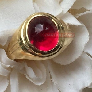 Peut inclure: Une bague en or avec une grande pierre précieuse rouge foncé sertie au centre. La bague a un design simple et classique.