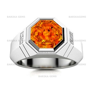 May include: A silver-toned ring featuring a large, octagonal-cut orange gemstone at the center. The band has a smooth, wide design with two small, rectangular-cut clear stones set into each side. The text BARIKA GEMS appears on the image.