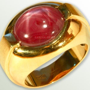 May include: A gold ring with a large, oval-shaped, red star ruby gemstone. The ruby has a star-like pattern in the center. The ring is set in a gold band with a smooth, rounded surface.