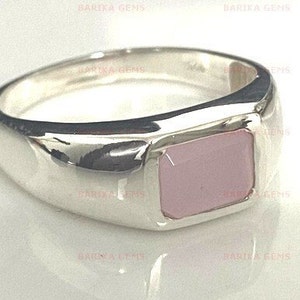 May include: A silver ring with a pink gemstone set in a square shape. The ring has a wide band and a polished finish.