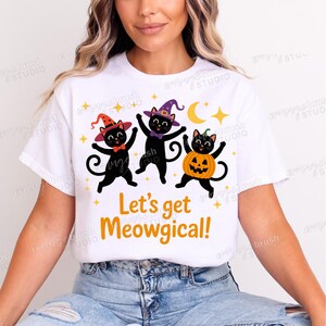 Puede incluir: Camiseta blanca con un dise&ntilde;o de Halloween. Presenta tres gatos negros con sombreros de bruja y un disfraz de calabaza. El texto "Let's get Meowgical!" est&aacute; impreso en naranja. El dise&ntilde;o incluye estrellas y una luna.