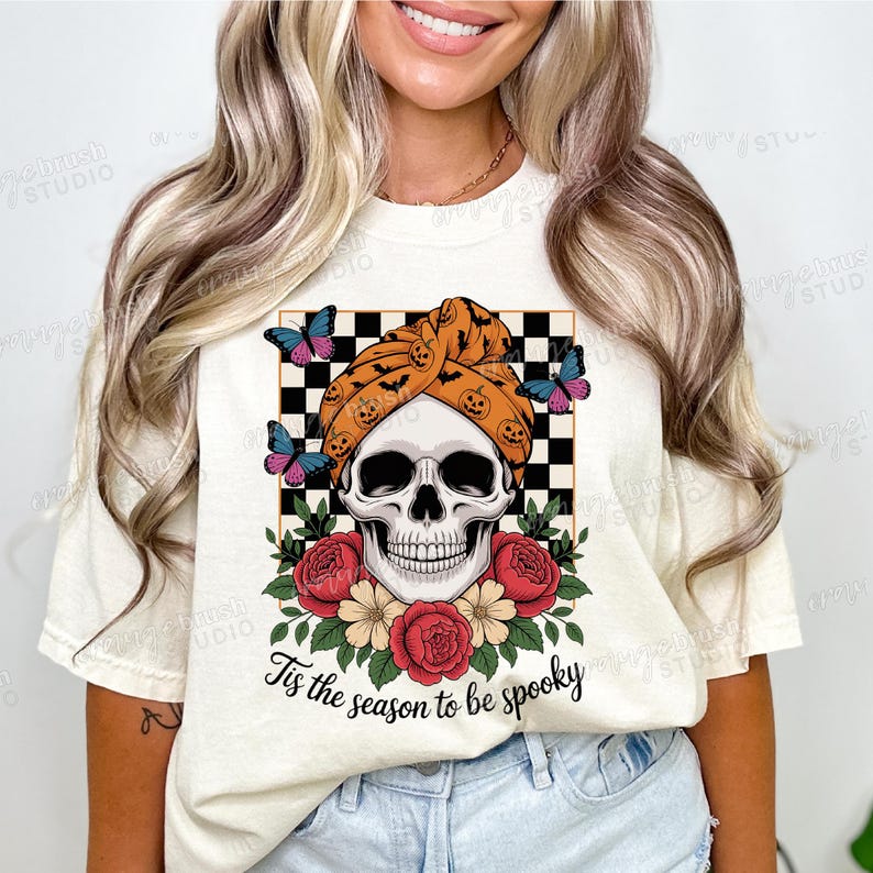 Puede incluir: Camiseta color crema con un dise&ntilde;o de Halloween. Presenta una calavera con un turbante con calabazas y murci&eacute;lagos, rodeada de rosas rojas y mariposas. Se incluye el texto "Tis the season to be spooky". El dise&ntilde;o est&aacute; sobre un fondo a cuadros blanco y negro.