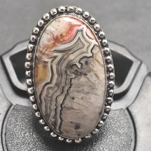 Op de afbeelding: Ovale zilveren ring met een grote, patroonsteen. De steen toont een mix van beige, grijze en rood-oranje wervelingen. De zilveren zetting heeft een kralenrand, wat het ontwerp van de ring versterkt.
