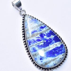 以下が含まれることがあります： 鮮やかな青と白の石が入った、涙型のペンダント。石は、ビーズの縁が付いた銀色の金属フレームにセットされています。ペンダントには、チェーンに取り付けるための銀色のベイルが付いています。