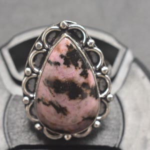 Op de afbeelding: Een zilveren ring met een druppelvormige edelsteen met roze en zwarte patronen. De steen is gezet in een decoratieve zilveren zetting met een geschulpte rand en kleine kraalaccenten. De ring wordt getoond op een grijze ondergrond.