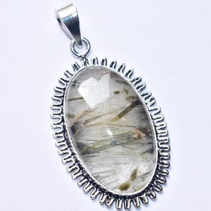 以下が含まれることがあります： 緑色のインクルージョンが入った透明な宝石が付いた楕円形のペンダント。銀色の金属フレームにセットされています。フレームは装飾的なスカラップエッジです。ペンダントにはチェーンを取り付けるためのシルバーベイルが付いています。