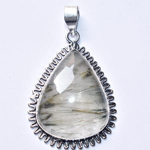 以下が含まれることがあります： 暗いインクルージョンを特徴とする、透明で明るい色の石が付いた涙の形のペンダント。 石は、装飾的なスカラップエッジを備えた銀色の金属フレームにセットされています。 ペンダントには、チェーン用のシンプルなベイルが付いています。