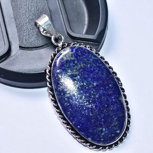 以下が含まれることがあります： 銀色のロープのような縁とベイルが付いた、楕円形のラピスラズリのペンダント。濃い青色の石には金色の斑点があり、背景に黒い物体がある白い表面に置かれています。 ジュエリー。