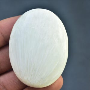 May include: An oval-shaped, off-white gemstone with a smooth, polished surface. The stone has subtle, light-colored veining throughout, giving it a natural, organic appearance. The gemstone is held in a hand against a dark background.