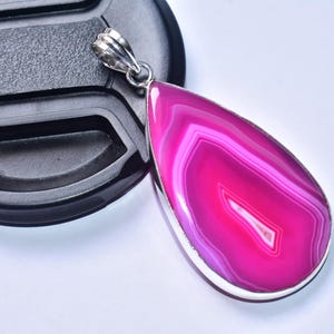 以下が含まれることがあります： ピンクと白のアゲートストーンを使用した、涙型のペンダント。石は銀色の金属フレームにセットされています。ペンダントは黒い表面に表示されています。