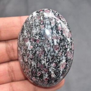 以下が含まれることがあります： 楕円形の研磨された石。 黒、白、赤紫色の斑点模様があり、独特の外観をしています。滑らかな表面は光を反射し、石の自然な質感を際立たせています。