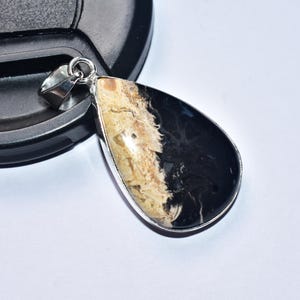 以下が含まれることがあります： 銀色のベイルが付いた、涙型のペンダント。ペンダントは、銀色のベゼルで囲まれた、研磨された黒とタンの石が特徴です。ペンダントは黒い表面と白い背景の上に置かれています。