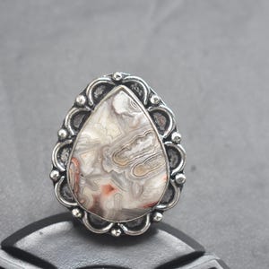 Op de afbeelding: Zilveren ring met een grote, druppelvormige steen met een uniek patroon van grijze, witte en roodbruine banden. De steen is gezet in een decoratieve, geschulpte bezel.