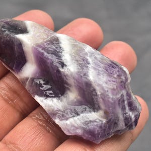 Könnte beinhalten: Ein rohes, unpoliertes Stück Chevron-Amethyst-Kristall mit ausgeprägten, abwechselnden Bändern aus tiefviolettem und undurchsichtigem weißem Quarz. Der Stein hat eine unregelmäßige, längliche Form mit einer rauen, texturierten Oberfläche.