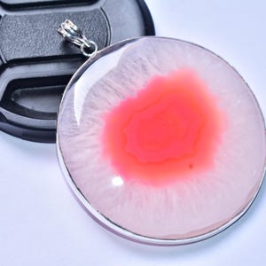 以下が含まれることがあります： 鮮やかな赤と白のアゲート模様が特徴の丸いペンダント。シルバーカラーのベゼルにセットされています。黒いカメラレンズキャップの隣に表示されており、ジュエリーやアクセサリーをテーマにしています。