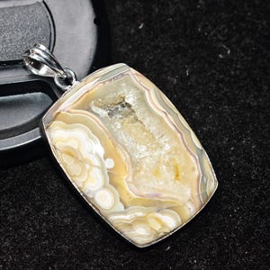 以下が含まれることがあります： 銀色のベイルが付いた長方形のペンダント。ペンダントは、ベージュ、クリーム、薄紫色の同心円状の帯を持つ研磨された瑪瑙石が特徴です。石は自然で有機的なパターンを持ち、銀色のベゼルにセットされています。