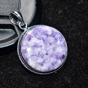 以下が含まれることがあります： 銀色のバチカンが付いた丸いペンダントで、質感のある紫色の石が特徴です。石には白と紫の大理石模様があります。ペンダントは、銀色のねじれたロープのような縁で囲まれています。背景は黒です。