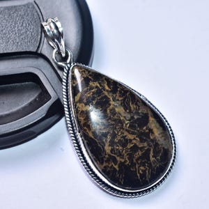 以下が含まれることがあります： ダークブラウンとゴールドの石が入った、涙型のペンダント。シルバーカラーの金属フレームにセットされています。ペンダントはテクスチャ加工された表面と、吊り下げ用のシルバーベイルが付いています。背景は白とグレーです。