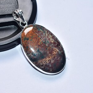 以下が含まれることがあります： 銀色の金属製のセッティングが付いた楕円形のペンダント。石は茶色、緑、タン色のまだら模様です。ペンダントは白い表面に表示されています。