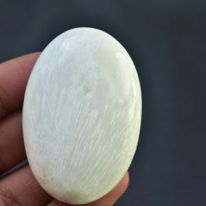 May include: A smooth, oval-shaped, off-white gemstone with subtle, natural veining. The polished surface reflects light, highlighting the stone's texture. The gemstone is held in a hand against a dark background.