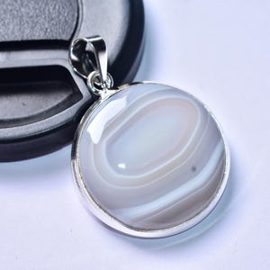 以下が含まれることがあります： シルバー色のベイルが付いた、丸いグレーと白のアゲートペンダント。ペンダントは、同心円状の色の帯を持つ研磨された表面を持っています。ペンダントはシルバー色のベゼルにセットされています。ペンダントは黒いオブジェクトの隣に表示されています。