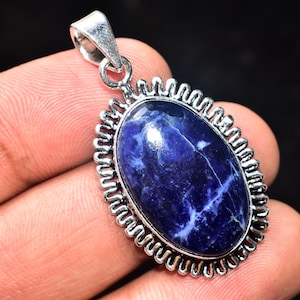 以下が含まれることがあります： 深い青色の石に白い縞模様が入った、銀色の金属フレームに収められた楕円形のペンダント。フレームは装飾的なスカラップエッジです。ペンダントは銀色のベイルから吊り下げられています。