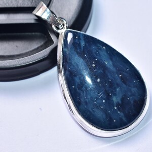 以下が含まれることがあります： 暗い青色のマーブル模様の石が入った、涙型のペンダント。石には白い斑点があり、銀色の金属フレームにセットされています。ペンダントは白い表面に置かれています。