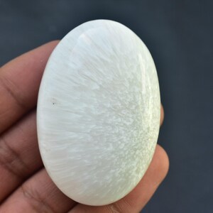 May include: A polished, oval-shaped white gemstone with a swirling, textured pattern. The stone is held in a hand against a dark background, showcasing its smooth surface and natural variations. The gemstone is approximately 2 inches in length.