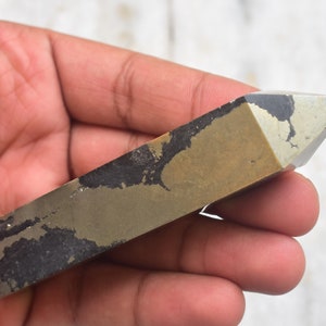 May include: A rectangular pyrite crystal with a pointed top. The crystal has a metallic gold color with dark gray and black patches. The crystal is held in a hand.