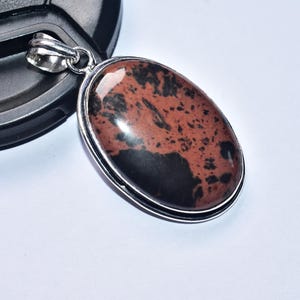 以下が含まれることがあります： 銀色の金属フレームにセットされた、磨かれた赤茶色と黒色の石の楕円形のペンダント。 ペンダントにはチェーン用のベイルがあり、白い表面に表示されています。