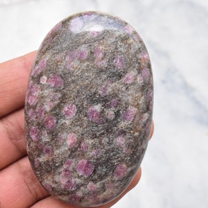 May include: An oval-shaped polished stone featuring a dark gray and brown matrix embedded with numerous vibrant pink, crystalline inclusions. The surface of the stone is smooth and reflective, showcasing a natural speckled pattern throughout the piece.