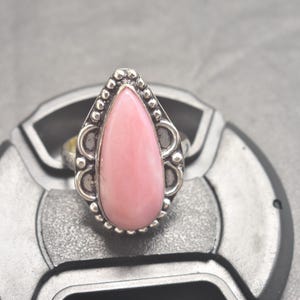 Op de afbeelding: Een zilveren ring met een druppelvormige, lichtroze edelsteen. De edelsteen is gezet in een gedetailleerde, sierlijke zilveren zetting met een kralenrand. De ring wordt getoond op een grijze ondergrond.