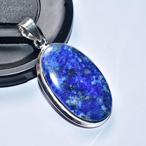 以下が含まれることがあります： 銀色のベイルが付いた、楕円形のラピスラズリペンダント。濃い青色の石には金の斑点があり、銀色のベゼルにセットされています。ペンダントは白い表面に表示されています。