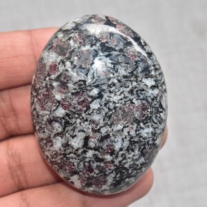 Może przedstawiać: Owalny, polerowany kamień szlachetny z nakrapianym wzorem czerni, bieli i głębokiej czerwieni. Powierzchnia odbija światło, podkreślając faktury i kolory kamienia. Unikalny przedmiot dekoracyjny.