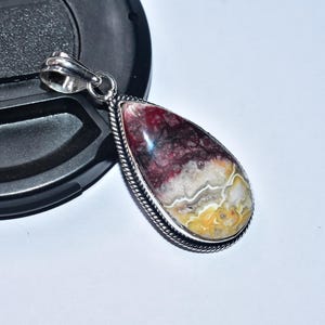 以下が含まれることがあります： 銀色のバチカンとセッティングが付いた、涙型のペンダント。石は、バーガンディ、白、黄色などの色の組み合わせが特徴です。ペンダントは白い背景と黒いオブジェクトの上に置かれています。