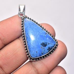 May include: A triangular pendant with a vibrant blue stone, accented with dark blue and black inclusions. The stone is set in a silver-colored metal frame with a beaded edge and a small loop for a chain.