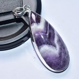 以下が含まれることがあります： 銀色の金属フレームにセットされた、磨かれた紫と白の石が付いた、涙の形をしたペンダント。石は大理石のような外観をしています。ペンダントは白い表面に表示されています。