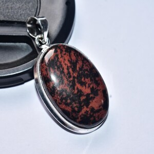 以下が含まれることがあります： 銀色の金属フレームにセットされた、黒い縞模様のある磨かれた赤褐色の石が付いた楕円形のペンダント。 ペンダントにはチェーン用のループがあり、白い背景に対して暗い表面に置かれています。 ジュエリーです。