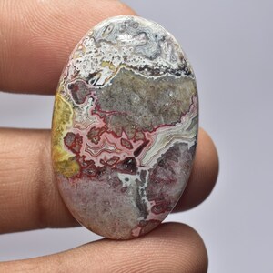 May include: An oval-shaped gemstone with a unique pattern of colors. The stone features a mix of white, gray, red, yellow, and brown hues, creating a marbled effect. The polished surface of the stone is smooth and reflective, showcasing the natural variations.