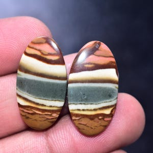 May include: Two oval-shaped gemstones with a polished finish. The stones display a layered pattern of colors, including shades of brown, cream, green, and pink. The gemstones are held in a person's hand.