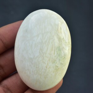 May include: Oval-shaped, polished gemstone with a creamy white base and subtle green and beige veining. The stone is held in a hand, showcasing its smooth, glossy surface. The gemstone's size is approximately 3 inches in length.