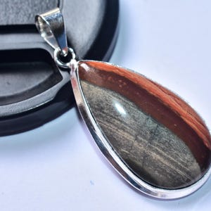 以下が含まれることがあります： 銀色のフレームが付いた、涙の形をしたペンダント。石は茶色、グレー、赤茶色の色合いです。ペンダントはネックレス用の銀色のベイルから吊り下げられています。