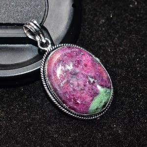 以下が含まれることがあります： 銀色の金属フレームにセットされた、大きな研磨されたルビーの宝石を特徴とする楕円形のペンダント。ルビーは濃いピンクと緑の色合いを示しています。ペンダントは黒い表面に表示されています。