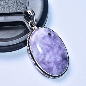 以下が含まれることがあります： 銀色の金属フレームにセットされた、大きく磨かれたラベンダー色の石の楕円形のペンダント。ペンダントには装飾的なロープのようなボーダーとチェーン用のシルバーベイルが付いています。背景は白です。