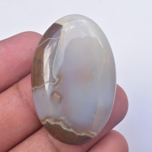 May include: Oval-shaped gemstone with a polished surface. The stone features a blend of colors, including brown, white, and gray, with natural patterns. The gemstone is smooth and appears to be a cabochon, suitable for jewelry making.