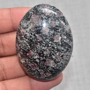 Könnte beinhalten: Ovaler Stein mit polierter Oberfläche. Der Stein ist überwiegend schwarz mit weißen und rot-violetten Einschlüssen. Die glatte, abgerundete Form deutet auf einen Handschmeichler oder ein dekoratives Objekt hin.